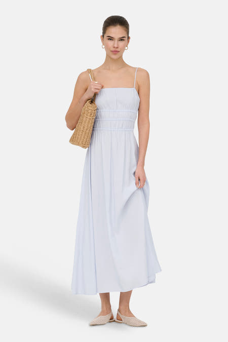 Robe longue bleu clair ONLY avec fines bretelles et taille smockée, stylisée avec un sac en osier et des chaussures plates beiges.