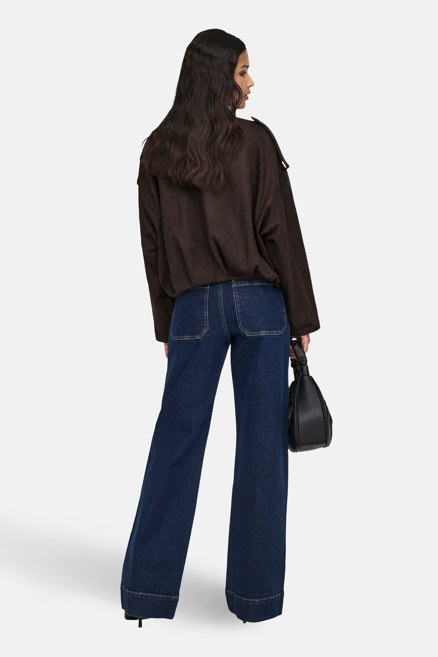 Een vrouw in een bruine blouse en wijde, donkerblauwe jeans, van achteren gezien, met een zwarte handtas.
