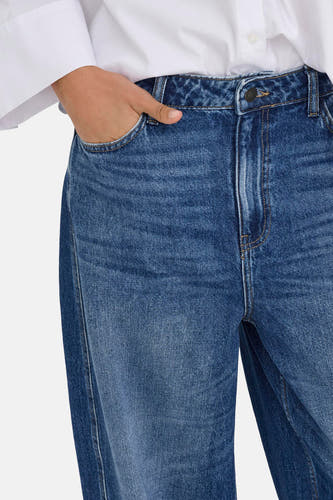 Jean en denim bleu marine spécial fit de JDY, avec une main dans la poche et une blouse blanche.