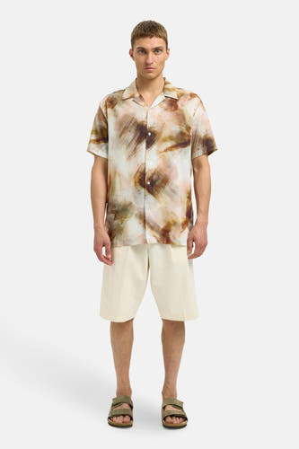 Chemise brune à manches courtes de SELECTED, avec un motif abstrait de traits de pinceau, portée avec des shorts crème et des sandales.