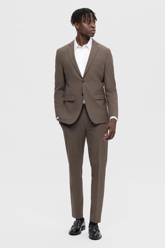 Man in bruin pak met wit overhemd en zwarte schoenen, staand tegen een witte achtergrond.