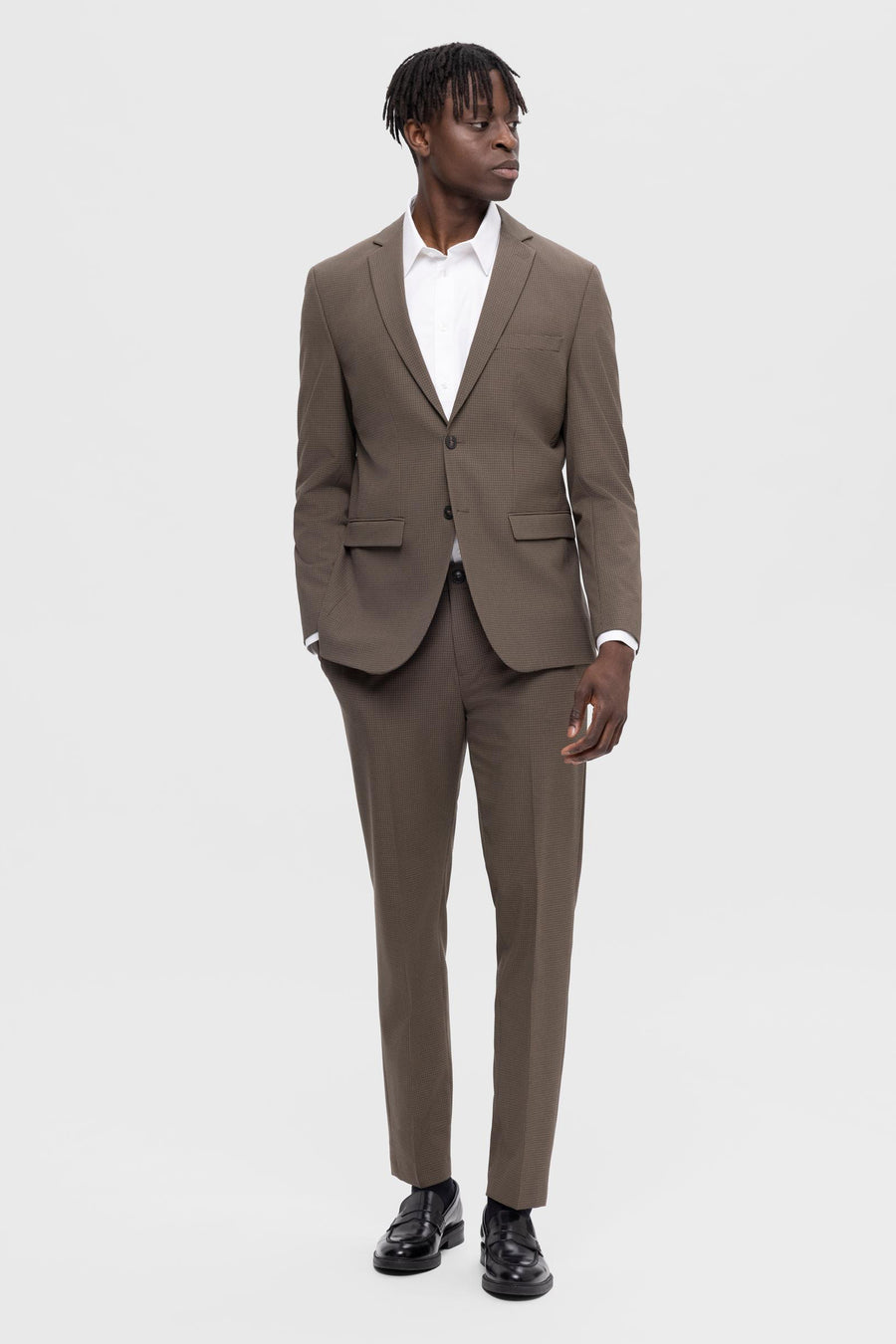 Man in bruin pak met wit overhemd en zwarte schoenen, staand tegen een witte achtergrond.