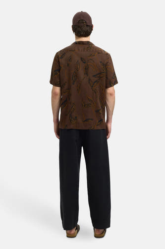 Chemise brune à manches courtes de SELECTED, avec un motif de feuilles, portée avec une casquette brune et un pantalon noir.