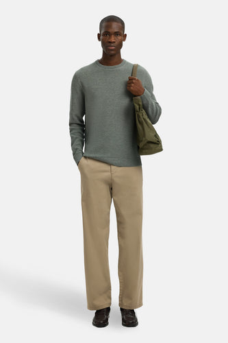 Man met groene trui, kaki broek en bruine schoenen, staand met tas.
