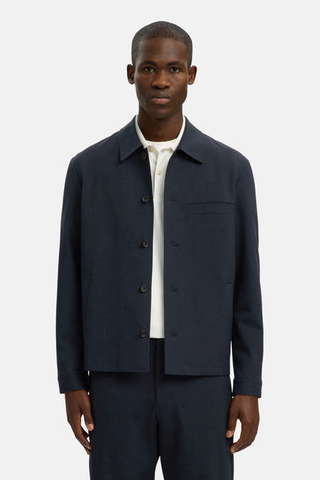 Chemise bleue à manches longues de SELECTED, avec une fermeture boutonnée et une poche poitrine, portée sur un polo blanc et un pantalon bleu foncé.