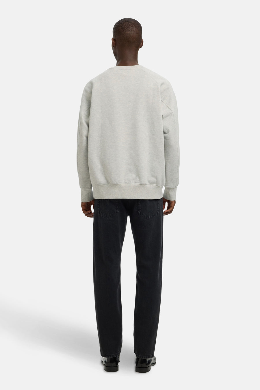 Achterkant van een man in grijze sweater, zwarte jeans en zwarte schoenen, staand tegen een witte achtergrond.