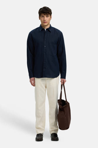 Chemise bleue à manches longues de SELECTED, portée par un homme avec un pantalon crème et un tote bag marron.