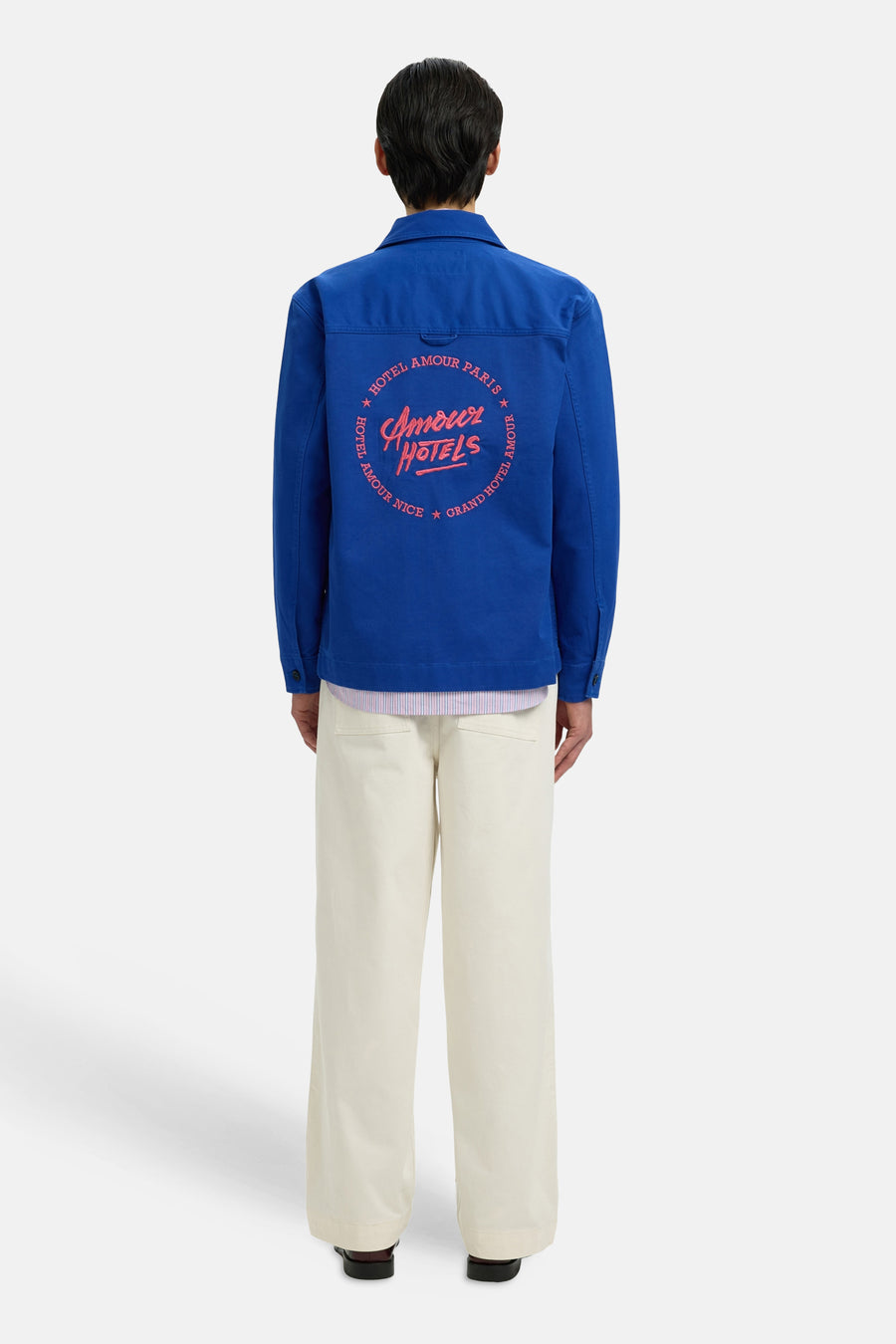 Veste bleue de SELECTED avec imprimé rose "Amour Hotels" au dos, portée avec une chemise rayée et un pantalon de couleur crème.