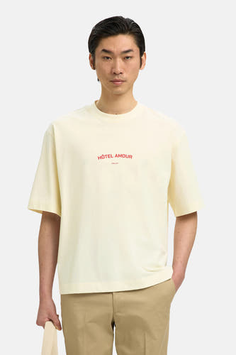 T-shirt écru de SELECTED, à manches courtes et avec le texte rouge "Hôtel Amour" sur la poitrine, porté avec un pantalon kaki.