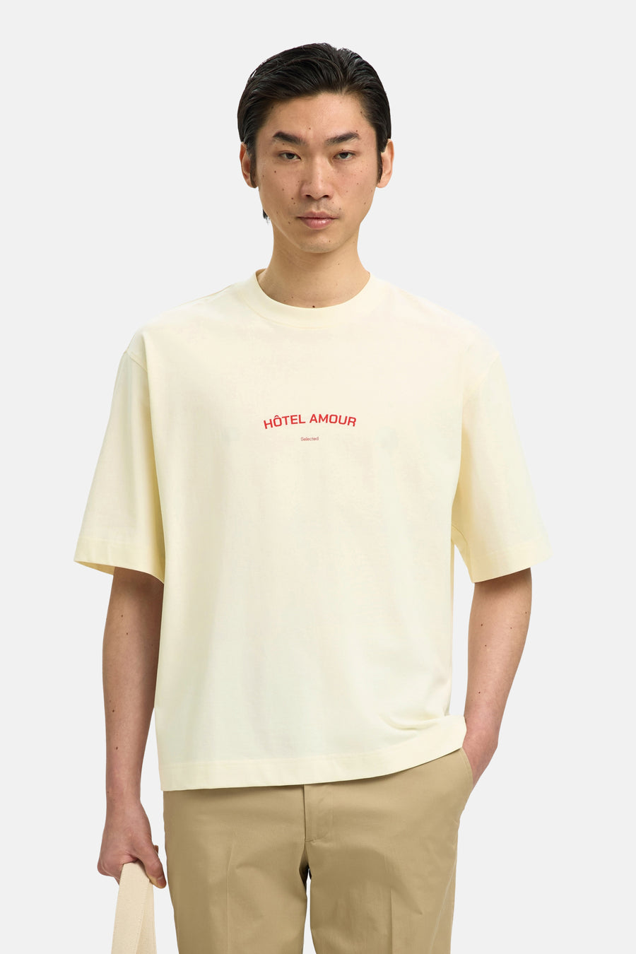 T-shirt écru de SELECTED, à manches courtes et avec le texte rouge "Hôtel Amour" sur la poitrine, porté avec un pantalon kaki.