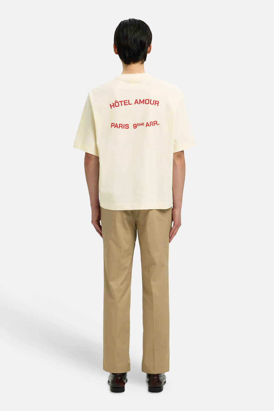 T-shirt écru de SELECTED avec texte rouge 'Hôtel Amour Paris 9ème Arr' au dos, porté avec un pantalon kaki et des chaussures marron.