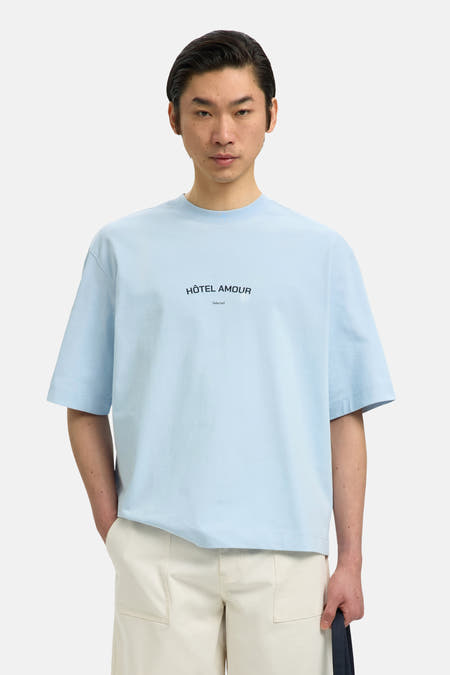 T-shirt bleu de SELECTED, à manches courtes et avec le texte "Hôtel Amour", porté avec un pantalon de couleur crème.