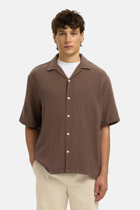 Chemise structurée marron à manches courtes de SELECTED, portée sur un t-shirt blanc, associée à un short beige et des sandales.