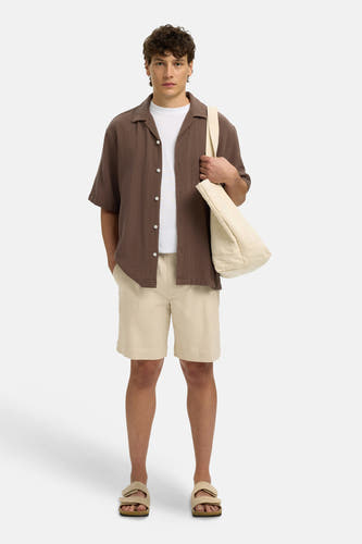 Chemise texturée marron à manches courtes de SELECTED avec boutons blancs, portée sur un t-shirt blanc, avec un short beige, des sandales et un sac fourre-tout.