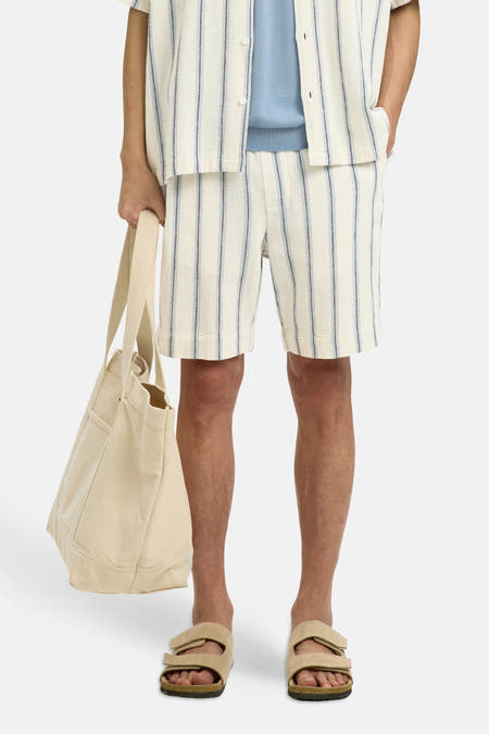 Ecru gestreepte korte broek van SELECTED, met fijne donkere strepen, lichte textuur, model in lifestyle pose met canvas tas en sandalen