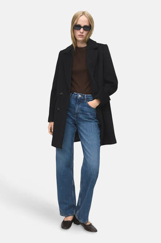 Manteau noir de MANGO, avec des manches longues, porté sur une chemise brune et un jeans bleu.