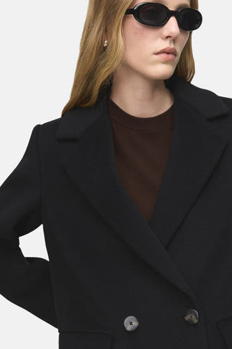 Manteau noir de MANGO, avec col revers et fermeture à boutons, associé à une chemise brune et à des lunettes de soleil noires.