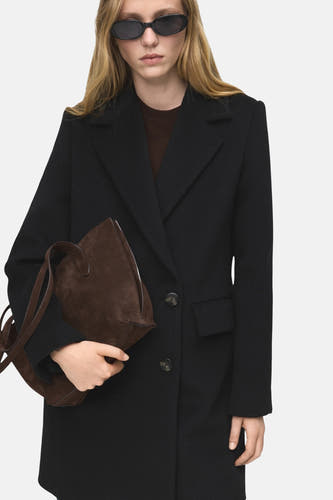 Manteau long noir de MANGO, porté avec une poche main brune et un chapeau de soleil ovale.