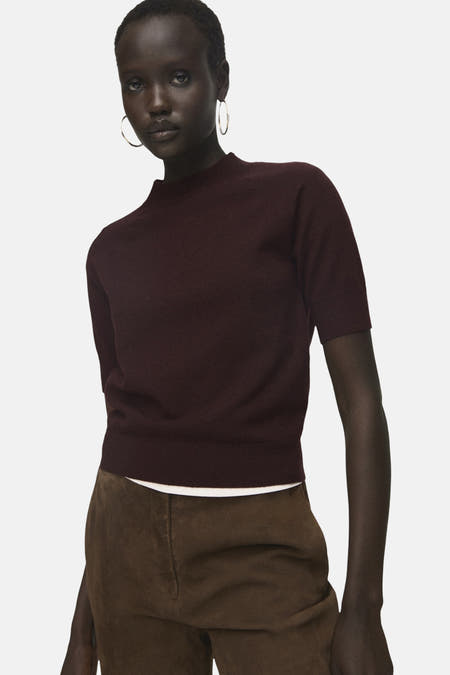 Veste Bordeaux avec col rond de MANGO, portée avec un pantalon marron et des boucles d'oreilles argentées.