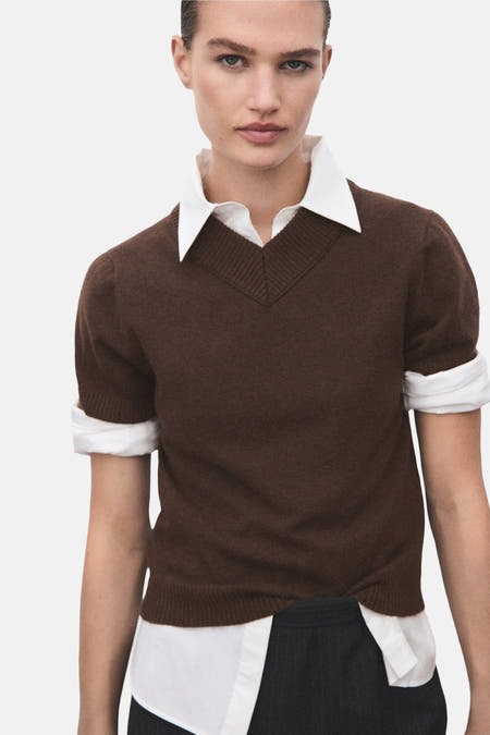 Pull marron avec col en V de MANGO, à manches courtes et une blouse blanche en dessous, associé à un pantalon foncé, vu de dos.