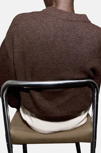 Sweat-shirt marron avec col rond de MANGO, vu de l'arrière, porté avec un t-shirt blanc et assis sur une chaise.