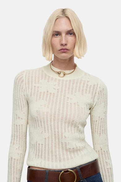 Pull beige à col rond de MANGO, avec un motif à rayures verticales et porté sur une jean bleu foncé avec une ceinture marron.