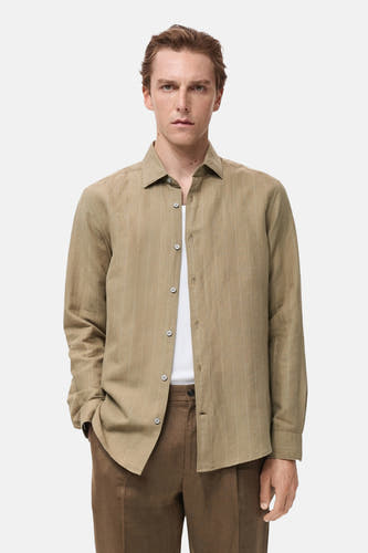 Chemise kaki à manches longues de MANGO, portée sur un t-shirt blanc, avec des pantalons marron et des sandales noires.