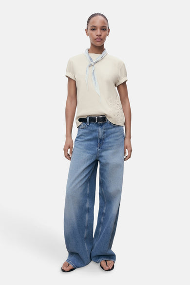 T-shirt beige à manches courtes de MANGO, avec des détails floraux et un foulard bleu autour du cou, associé à une ceinture noire et des jeans larges.