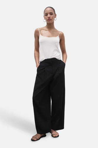 Pantalon de costume noir de MANGO, porté avec un chemisier blanc et des sandales, les mains dans les poches.