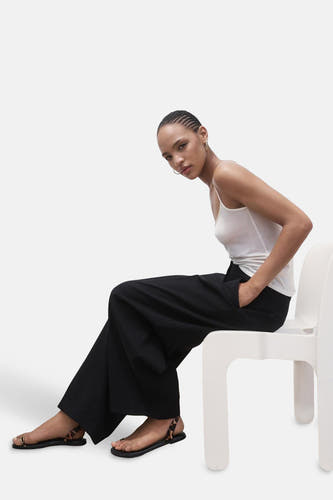 Pantalon de costume noir MANGO, porté avec une chemise blanche et des sandales à imprimé léopard, assis sur une chaise blanche.