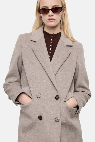 Manteau long marron de MANGO, avec une fermeture à double boutons et des poches, porté sur un chemisier marron à colliers.
