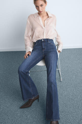 Jean flare bleu marine de MANGO, porté avec un chemisier rayé et des escarpins marrons, assis sur une chaise.
