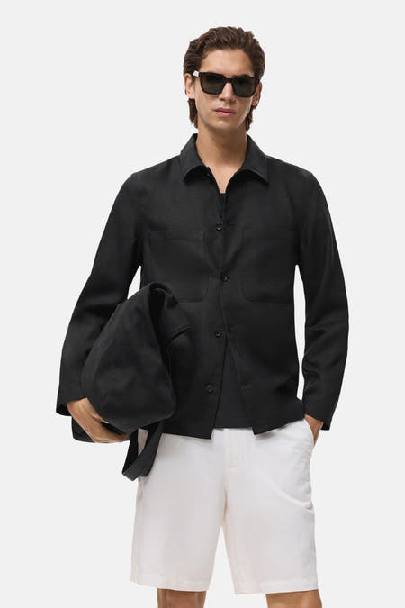 Manteau court noir de MANGO, avec des manches longues et des poches, porté avec un short blanc et un chapeau de soleil.