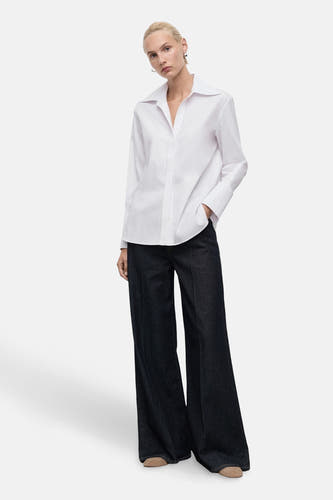 Chemise blanche à manches longues de MANGO, portée avec un pantalon large et sombre et des chaussures.