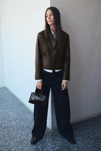 Vrouw in bruine leren jas, witte blouse, wijde donkere jeans en zwarte tas staand bij een witte muur.