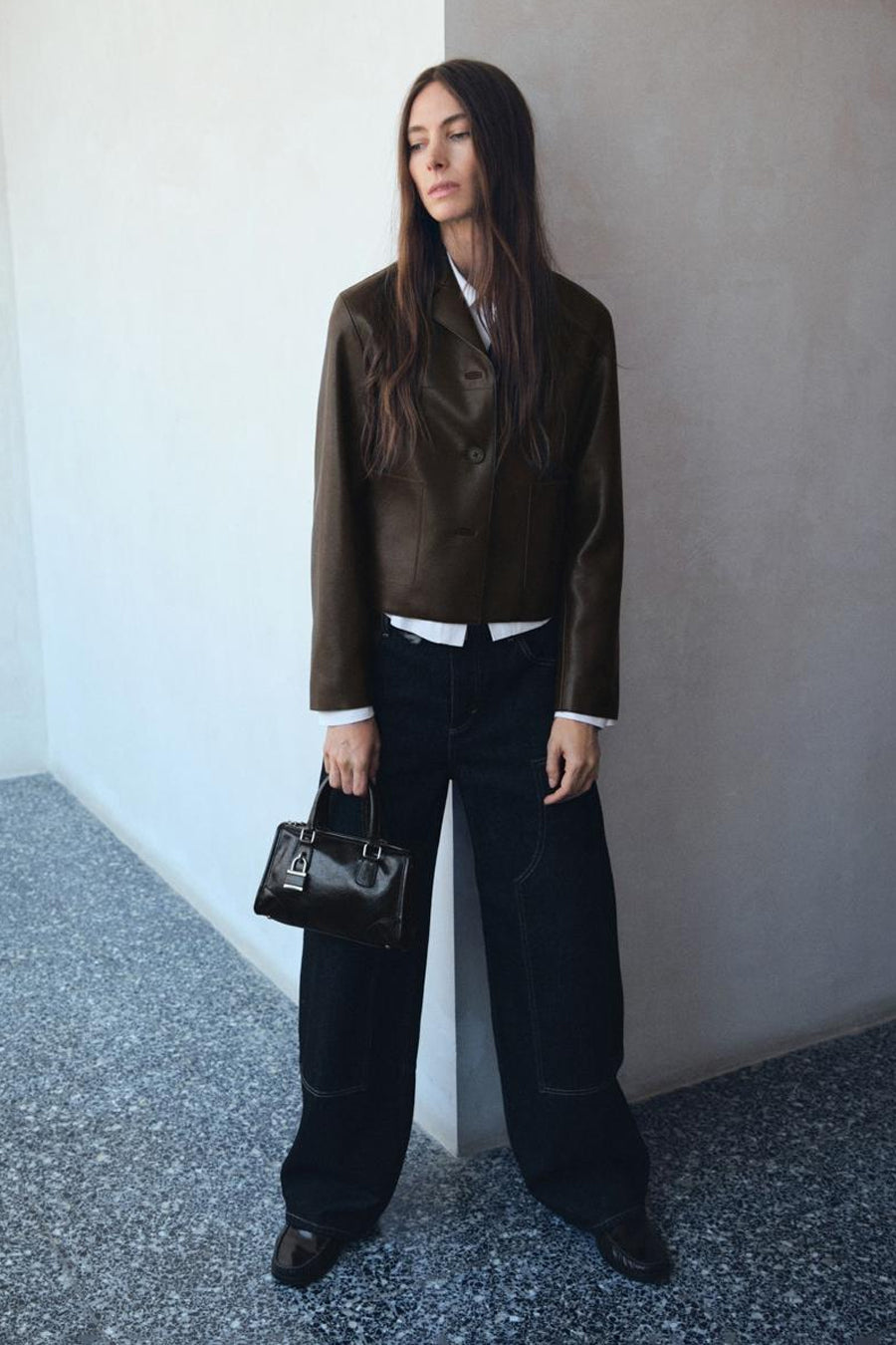Vrouw in bruine leren jas, witte blouse, wijde donkere jeans en zwarte tas staand bij een witte muur.