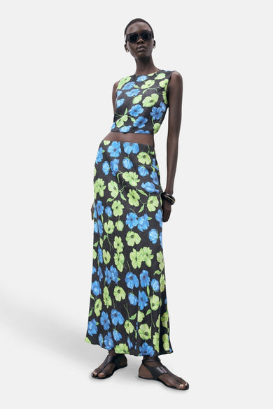 Long jupe multicolore de MANGO, avec un imprimé fleuri et portée avec des sandales noires et un chapeau de soleil.