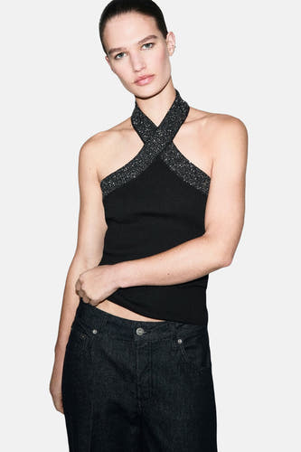 Blouse sans manches noire de MANGO, avec un bord pailleté, associée à des jeans foncés et une queue de cheval.