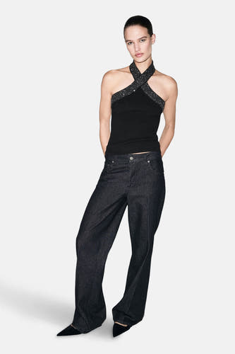 Zwarte mouwloze blouse van MANGO, met gekruiste glitterbanden en gecombineerd met wijde donkerblauwe jeans en zwarte puntige pumps.
