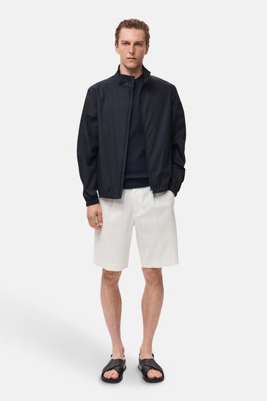Manteau bleu court de MANGO, avec une fermeture éclair, porté avec un pull bleu marine et des short rayés.