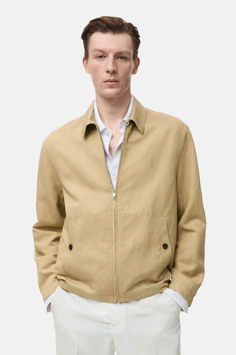 Manteau court beige de MANGO, avec fermeture éclair et poches, porté sur une chemise blanche et un pantalon blanc.