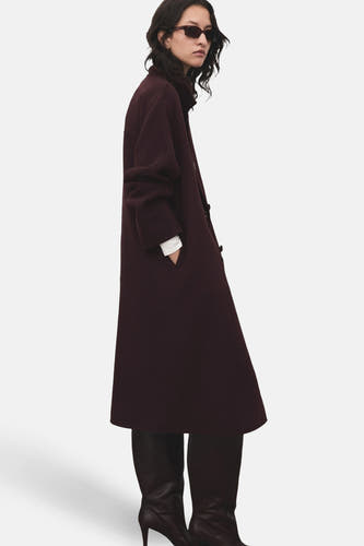 Manteau long de MANGO, avec poches, associé à des bottes noires à talon et à un masque solaire.