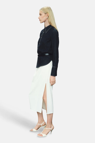 Chemise en denim bleu marine à manches longues de MANGO, associée à une jupe blanche avec fente et talons blancs.
