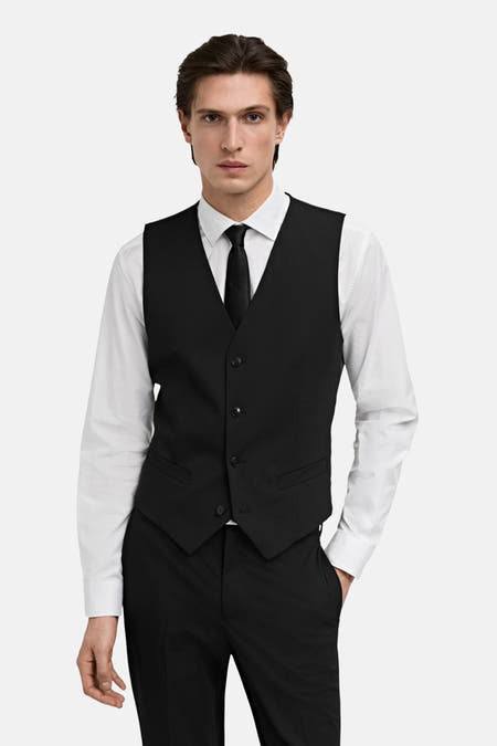Gilet noir de MANGO, porté sur une chemise blanche avec une cravate noire et un pantalon noire.