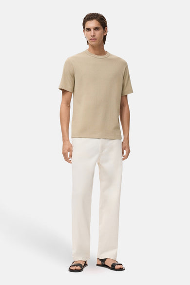 T-shirt beige à manches courtes de MANGO, avec une texture lisse, porté avec un pantalon blanc et des sandales noires.