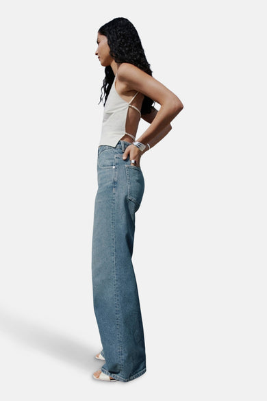 Jean en denim bleu mid, large de MANGO, avec des jambes larges et un design à cinq poches, porté avec une chemise blanche et des sandales.