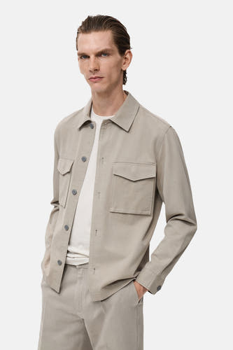 Chemise beige à manches longues de MANGO, avec poches et boutons, portée sur un T-shirt blanc et une pantalon beige.