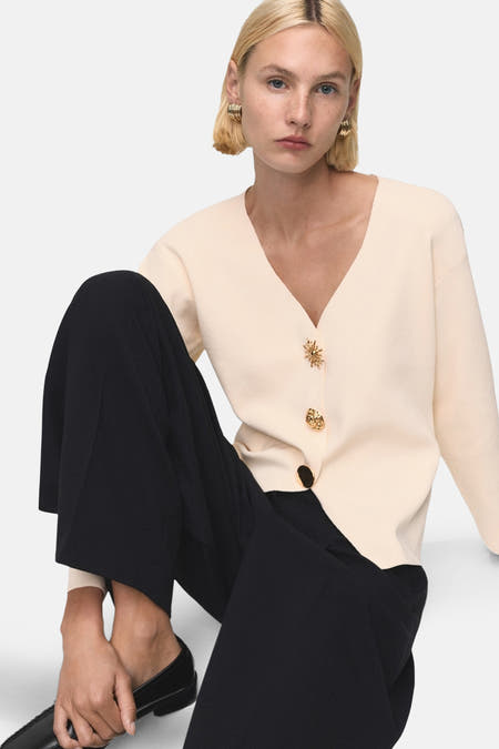 Gilet écru de MANGO avec des boutons décoratifs, associé à un pantalon noir et des chaussures noires.