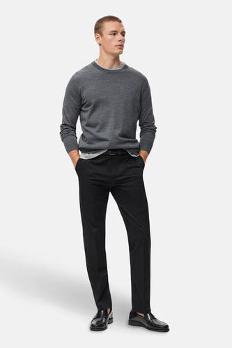 Pantalon bleu marine de MANGO, porté avec un pull gris et des chaussures en cuir noires, les mains dans les poches.