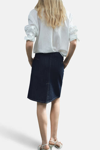 Jupe en denim bleu marine de MANGO, avec un zip asymétrique et des poches arrière, portée avec une chemise rayée et des sandales blanches.